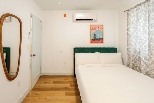 A hotel room with a wooden floor, a bed with white bedding, a green headboard, a wall-mounted air conditioner, a framed picture of Japan, and a patterned curtain.