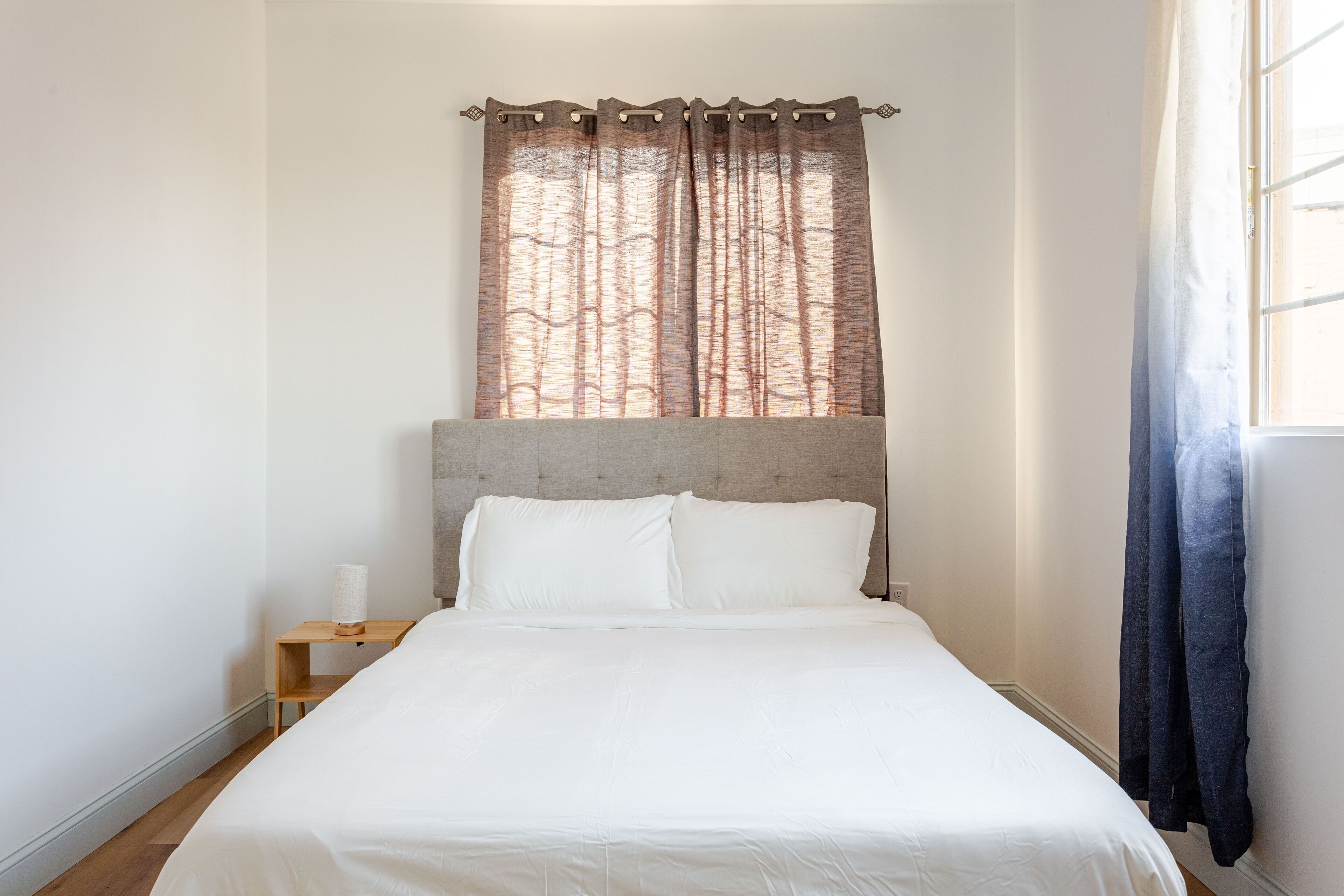A neatly made bed with white linens, a wooden nightstand with a lamp, and a window with a curtain.