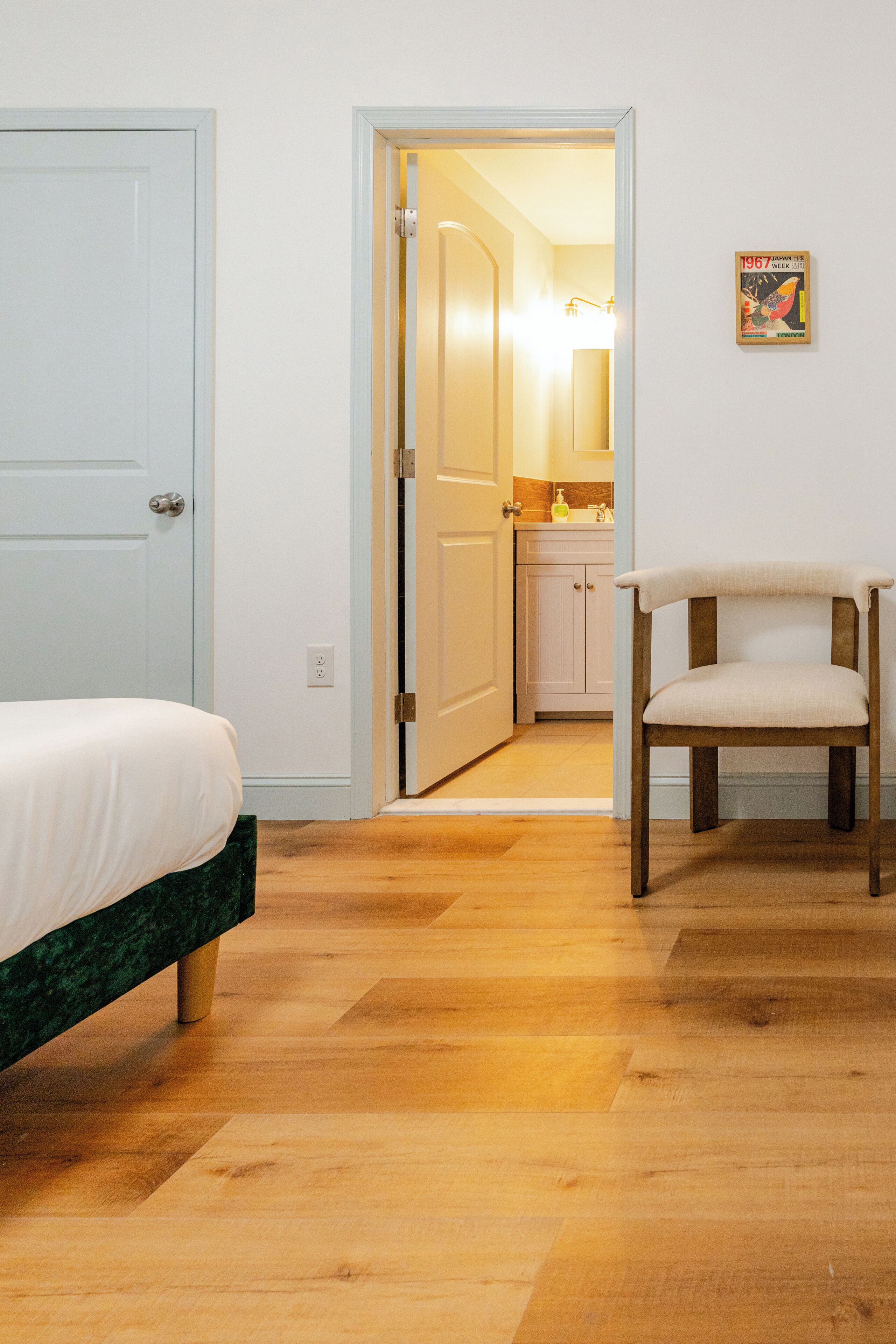 Un dormitorio con una cama, suelo de madera, una silla y un baño visible a través de una puerta abierta.