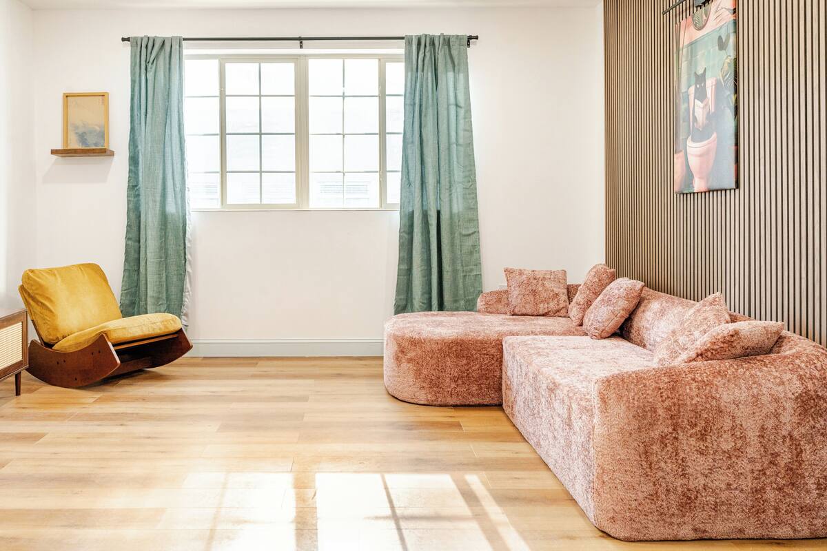 A living room with a wooden floor, a brown sofa, a yellow chair, and a window with curtains.