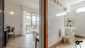 Una habitación de hotel moderna con balcón, escritorio con silla, ventana grande y baño con lavamanos y espejo.