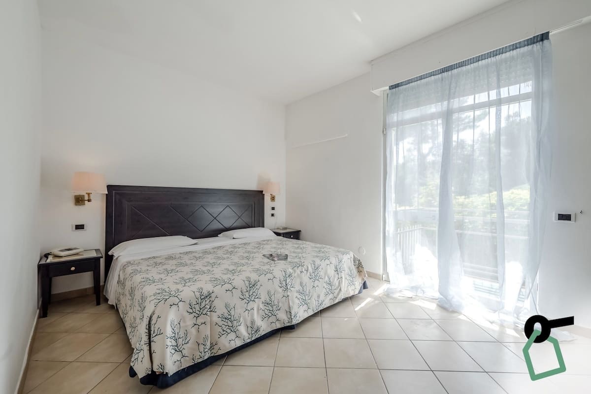 Quarto com cama, duas mesinhas de cabeceira, um criado-mudo, janela com cortinas finas e vista para o verde lá fora.