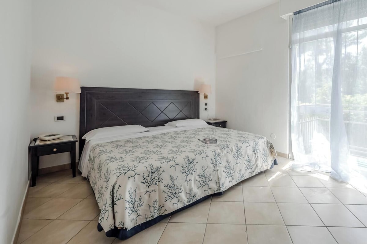 Une chambre à coucher comprenant un lit, deux tables de chevet, une commode, une fenêtre avec des rideaux légers et une vue sur la verdure à l’extérieur.