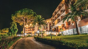 Un hotel bien iluminado con varios pisos, palmeras en el exterior y un camino pavimentado que lleva a la entrada.
