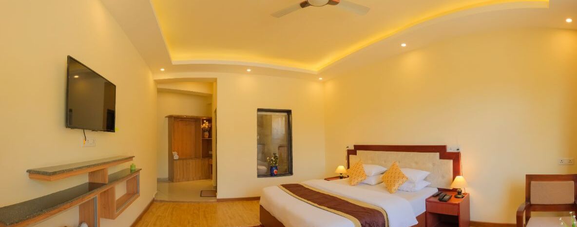 Quarto de hotel com cama, mesa de cabeceira, televisão e uma pequena janela.
