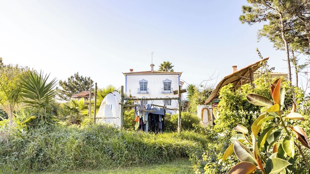 Un edificio blanco con tejado de baldosas, rodeado de abundante vegetación y una pequeña construcción con techo de paja.