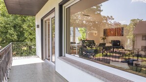 Ein Balkon mit Metallgeländer, eine Glastür zu einem Sitzbereich im Freien sowie ein Blick auf einen Garten und parkende Autos.