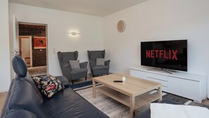 Ein modernes Wohnzimmer mit einem schwarzen Ledersofa, zwei grauen Sessel, einem Holztisch und einem Fernseher, der "NETFLIX" anzeigt.