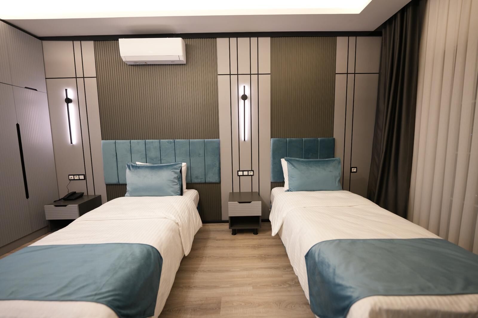 Una habitación de hotel moderna con dos camas, aire acondicionado integrado, mesitas de noche y un gran ventanal con cortinas.