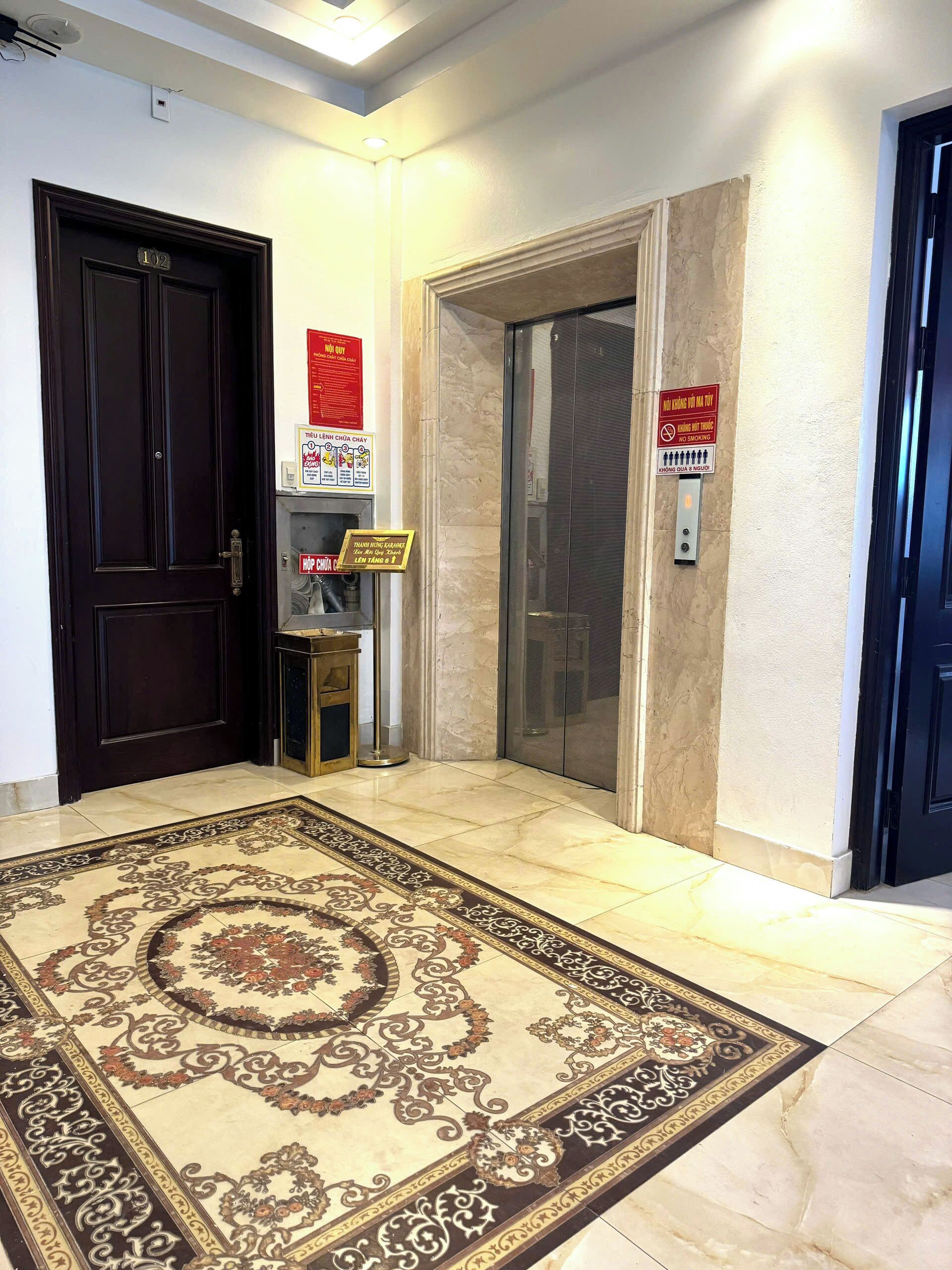 A lobby with a dark wooden door, a marble floor, and an elevator. There are signs on the wall, including one that says "No smoking."
