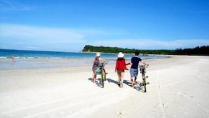 A sandy beach with three people riding bicycles, a clear blue sky, and a distant shoreline with greenery. - Paloma Hotel (Van Don)
