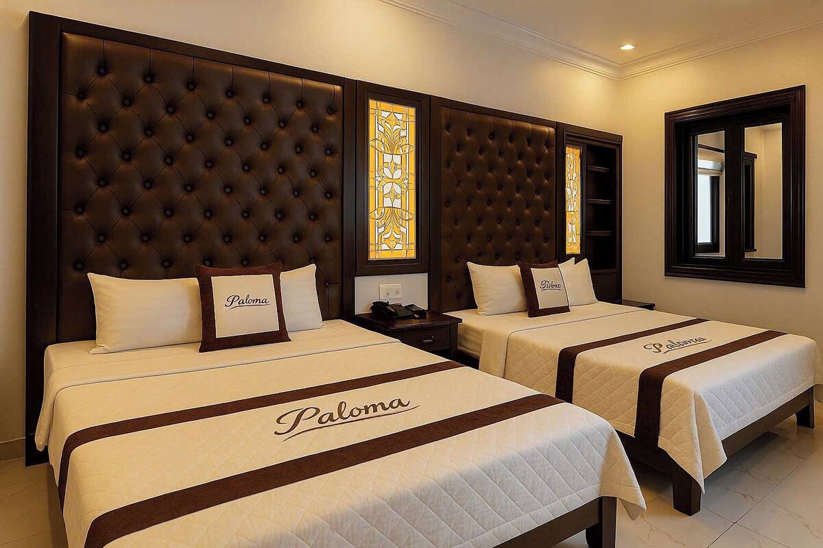 A hotel room with two beds, each with a headboard and a bedspread with "Paloma" embroidered on it.