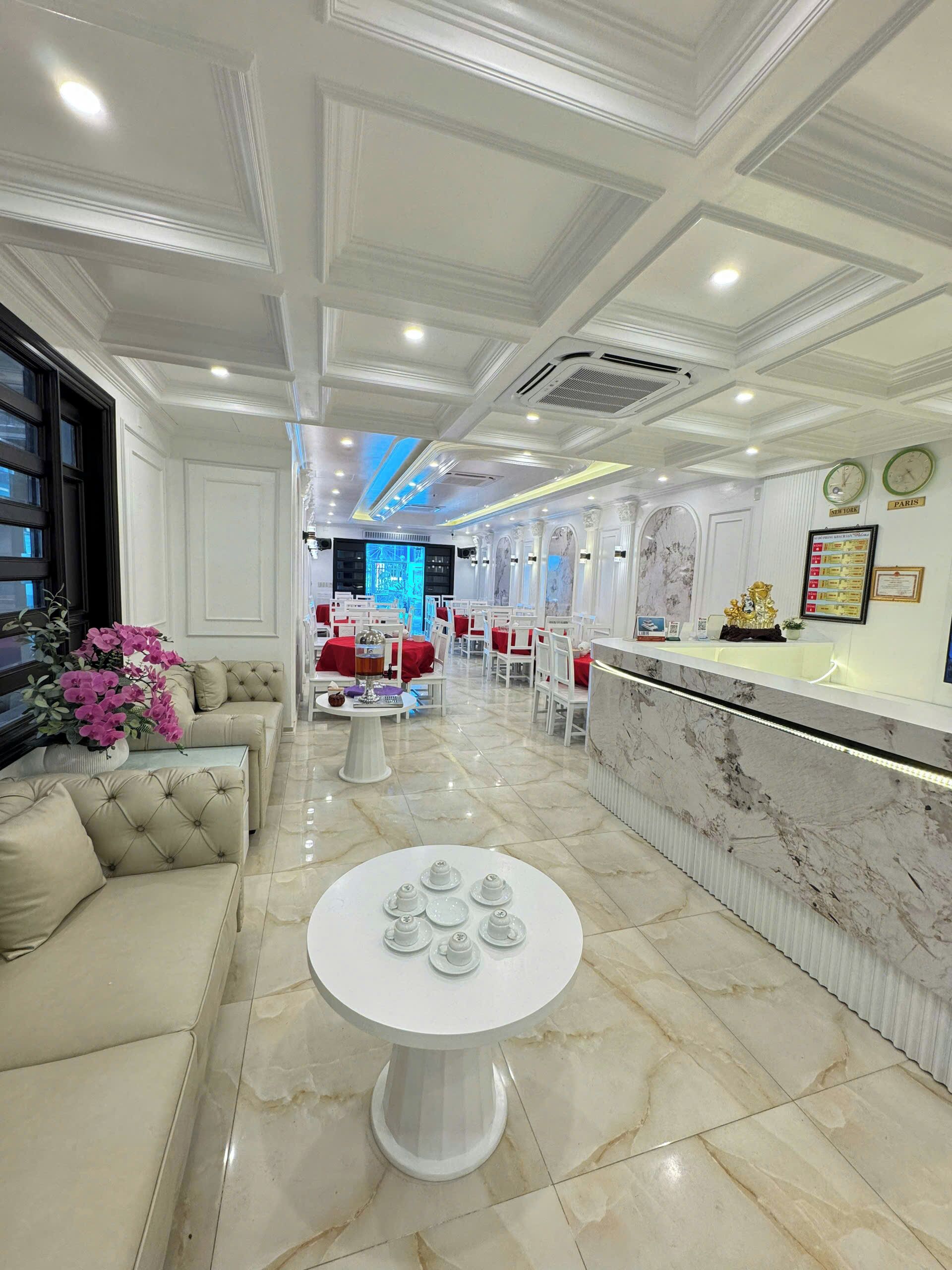 A hotel lobby with a marble reception desk, a seating area with sofas and a round table, and a decorative wall with clocks and framed certificates.