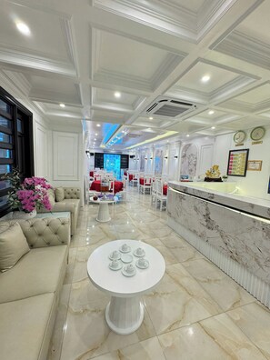 A hotel lobby with a marble reception desk, a seating area with sofas and a round table, and a decorative wall with clocks and framed certificates.