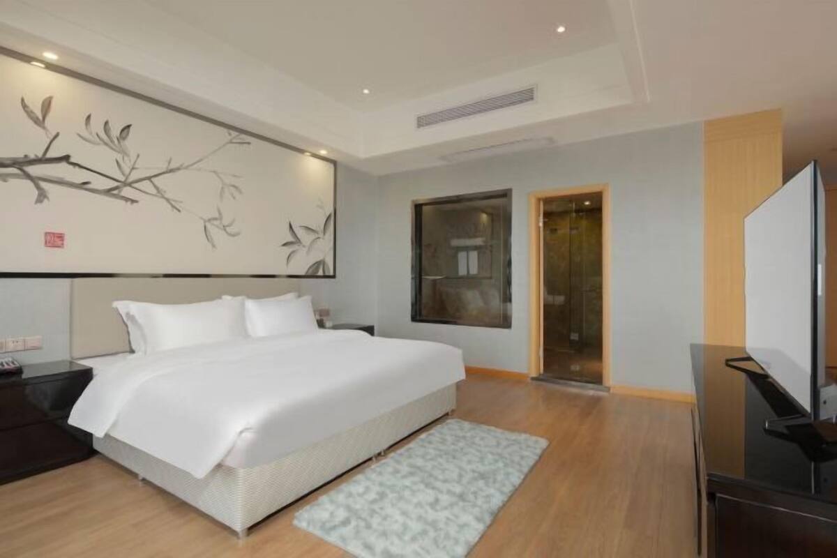 A modern hotel room with a large bed, a flat-screen TV, a wall-mounted artwork, and a sliding door leading to a bathroom.