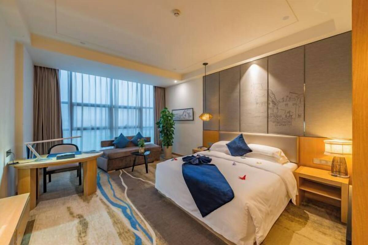 A hotel room with a bed, a desk, a chair, a sofa, a lamp, and a window with curtains.