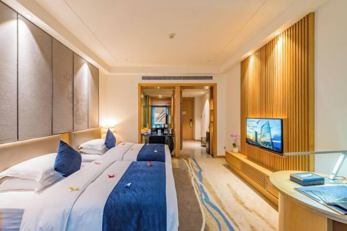 A hotel room with a large bed, a TV mounted on a wooden panel, and a desk with a lamp.