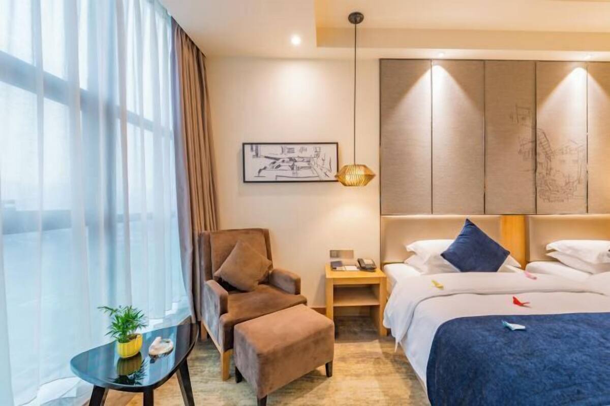 A hotel room with a bed, a chair, a small table with a plant, and a wall-mounted artwork.