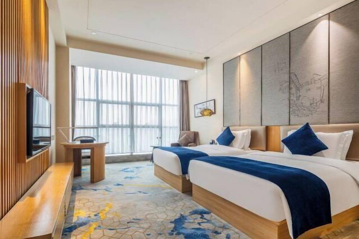 A hotel room with two beds, a TV, a desk, and a large window with blinds.