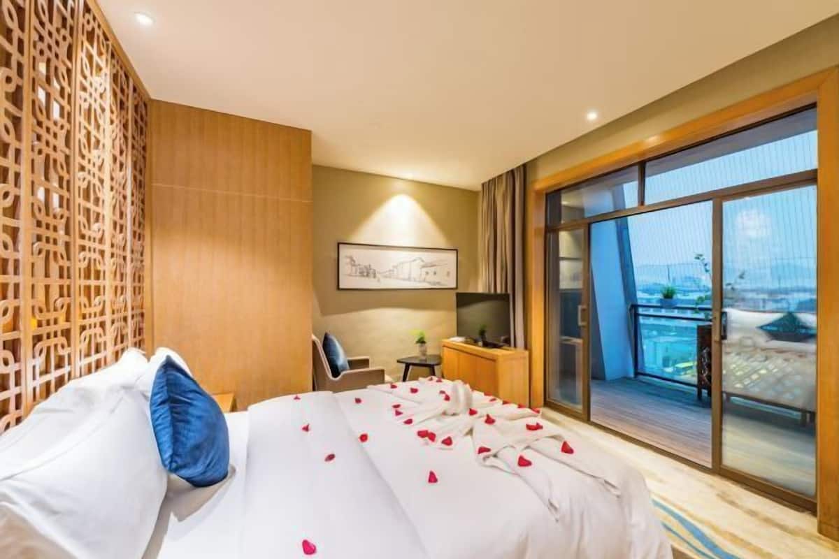 A hotel room with a bed, a desk, a chair, a small table with a plant, and a large window with a view of the city.