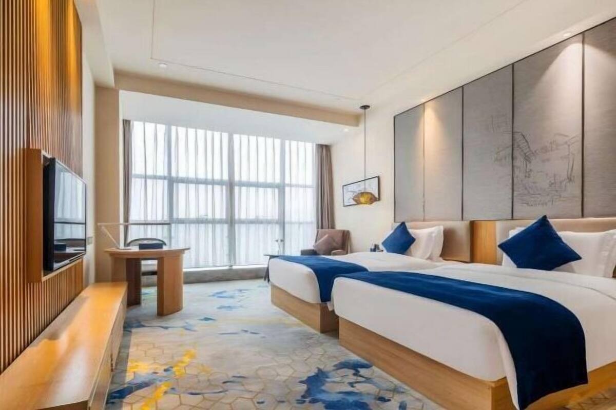 A hotel room with two beds, a TV, a bench, and a large window.