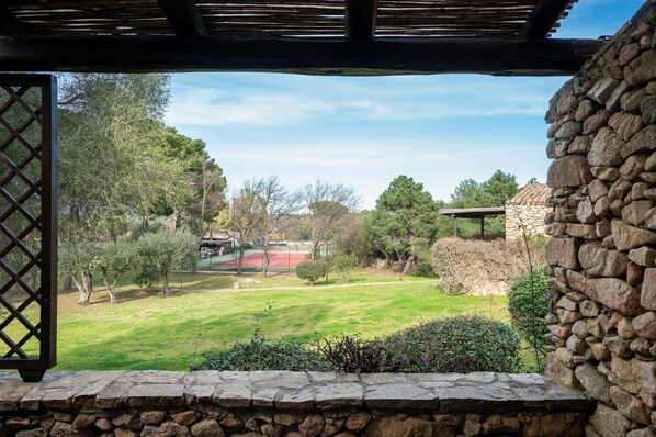 A stone wall and a wooden pergola overlooking a well-maintained garden with a tennis court.
