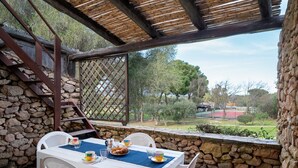 A covered patio with a stone wall, a table with chairs, and a view of a park and buildings in the distance. - Caletta Nest 38 by Klodge – Pool & tennis (Porto Rotondo)