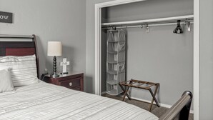 A bedroom with a bed, bedside table, lamp, and a wardrobe with a hanging shelf. - Clean, Cozy 2BR, Patio, Near Razorback Stadium (Springdale)
