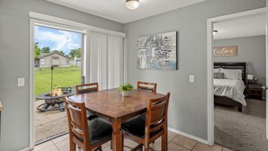 A dining area with a wooden table and chairs, a view of a backyard, and a bedroom with a bed and nightstand. - Clean, Cozy 2BR, Patio, Near Razorback Stadium (Springdale)