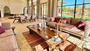 A spacious living room with a large window, a wooden coffee table, a sofa, and a dining area with chairs. - Palais Kenza (Marrakech)