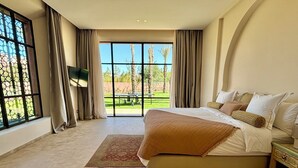 A spacious bedroom with a large bed, a TV, and a view of a garden through large windows. - Palais Kenza (Marrakech)