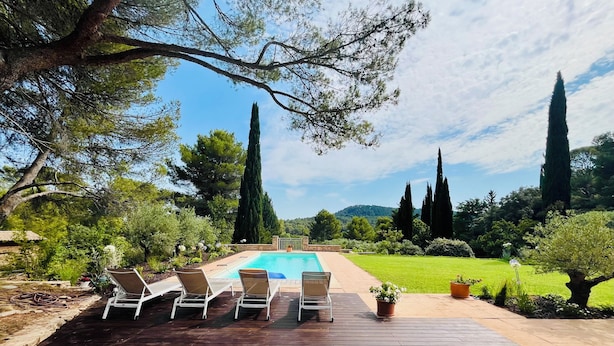 Una terraza de madera con sillones de descanso y una piscina, rodeada de árboles y césped.