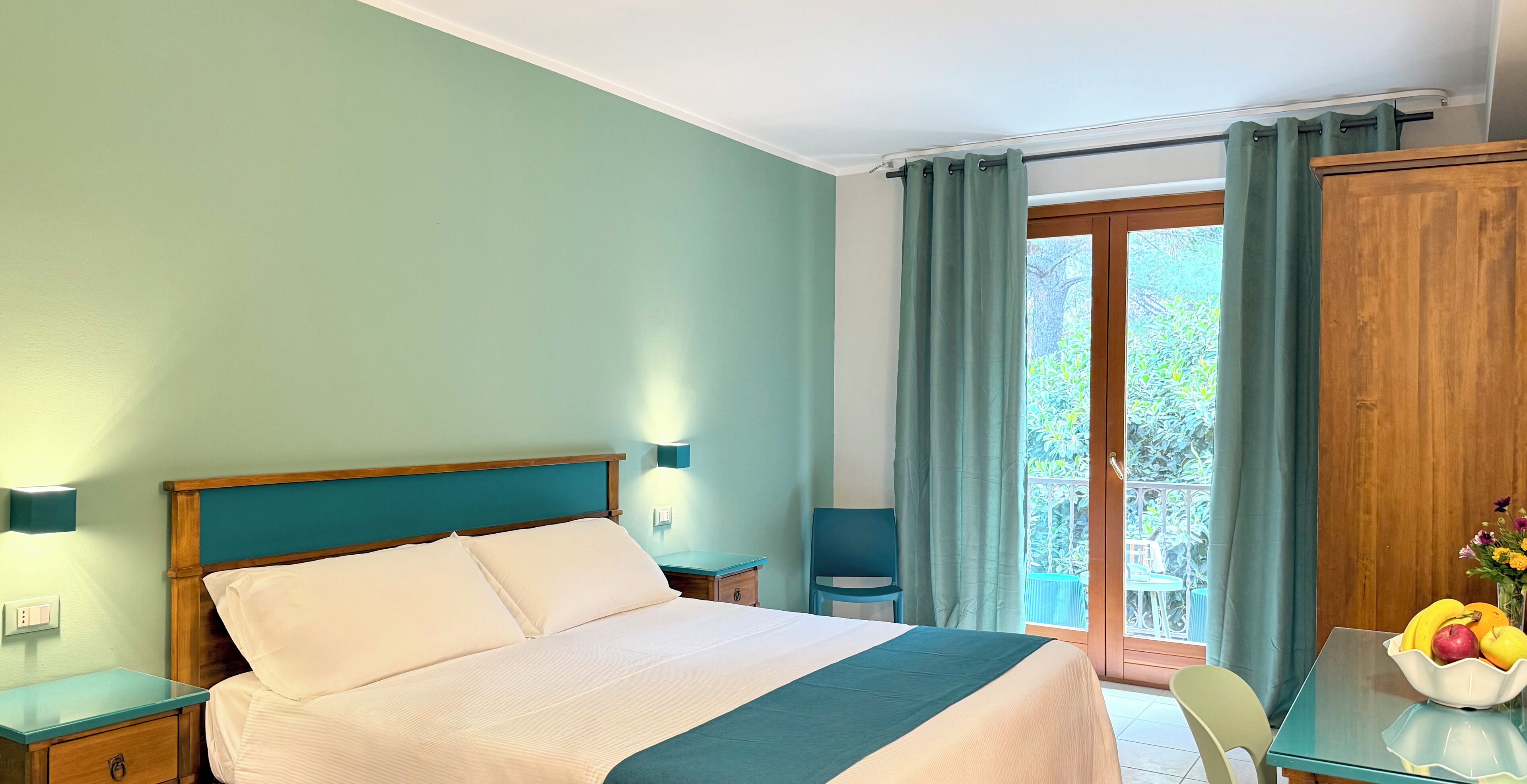 Una habitación de hotel con una cama, una mesita de noche, una mesa con frutero y vistas al exterior.