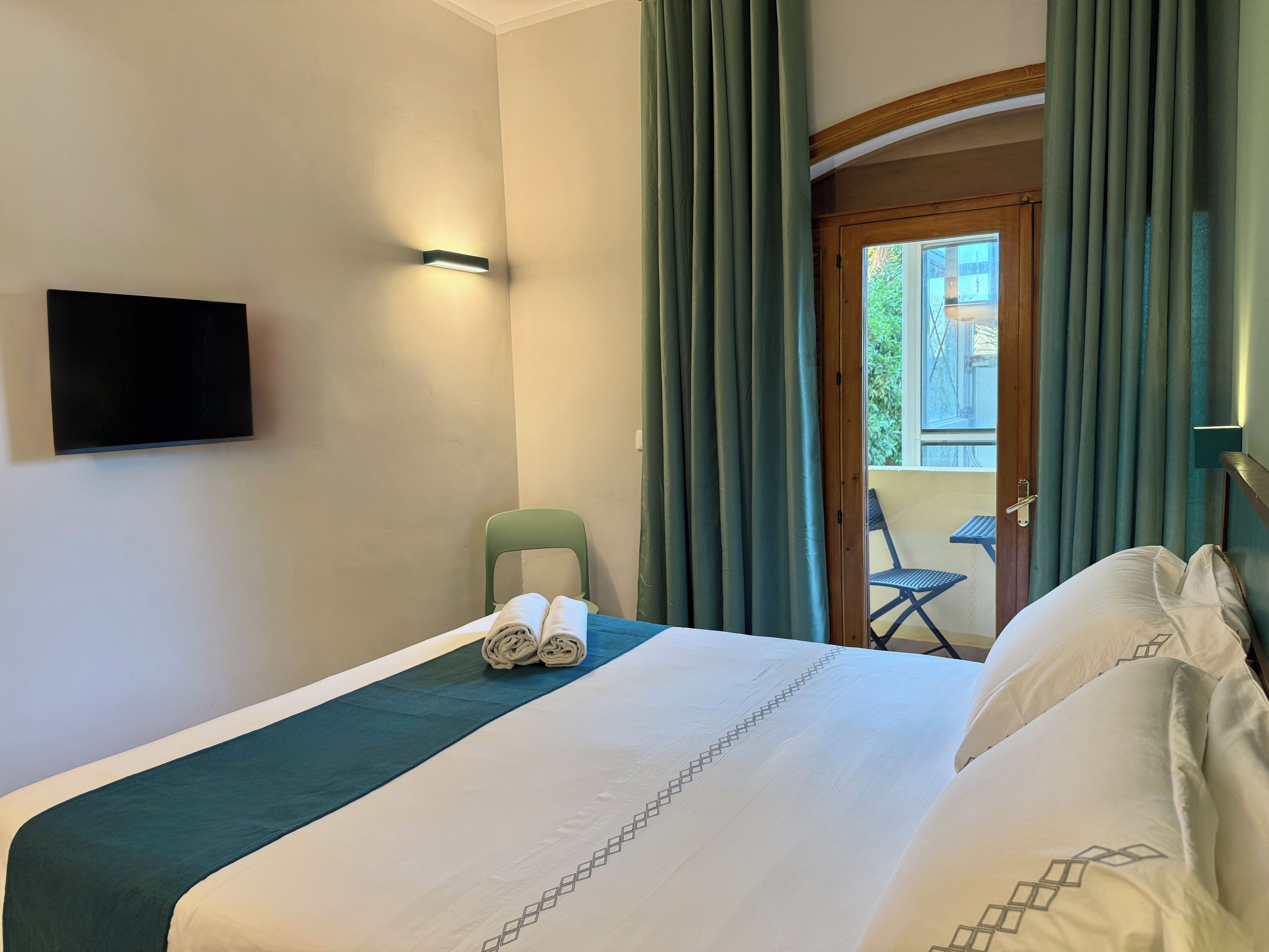 Ein Hotelzimmer mit einem Bett, einem an der Wand befestigten Flachbildfernseher, einem grünen Sessel und Blick auf den Balkon durch eine Tür.