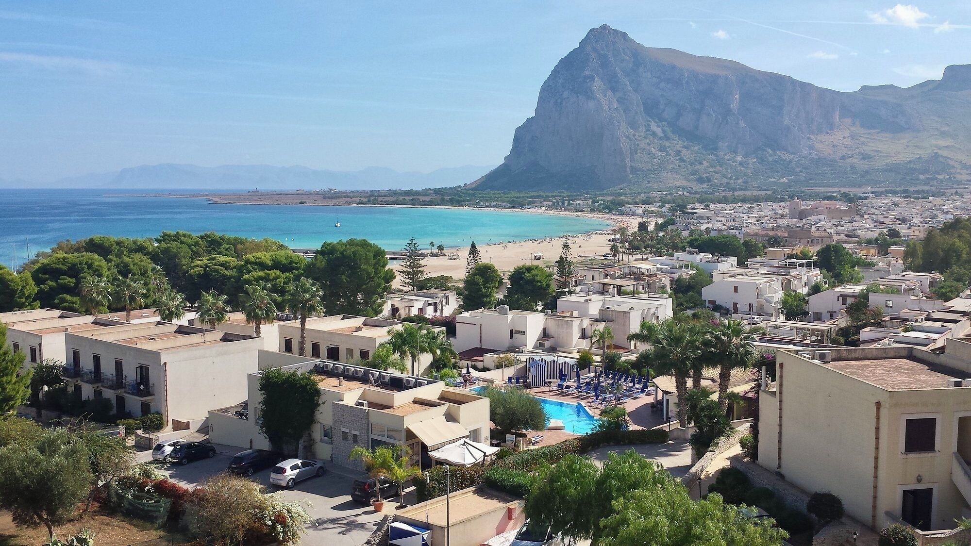 Une ville côtière avec une grande montagne en arrière-plan, une plage et un espace piscine doté de chaises longues.