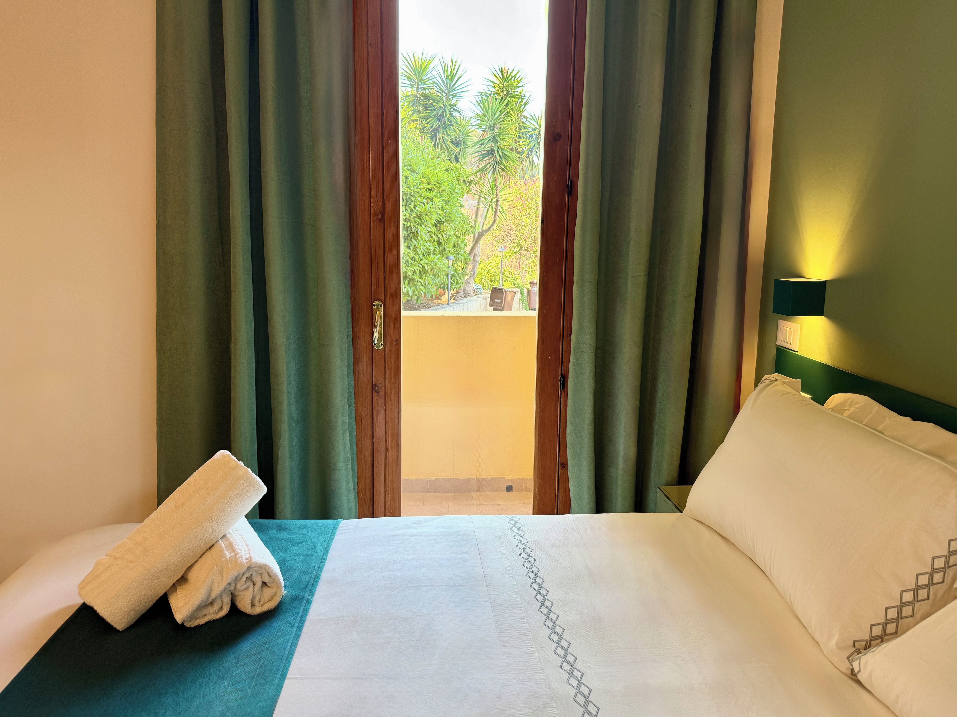 A hotel room with a bed, a view of greenery outside, and a towel on the side table.