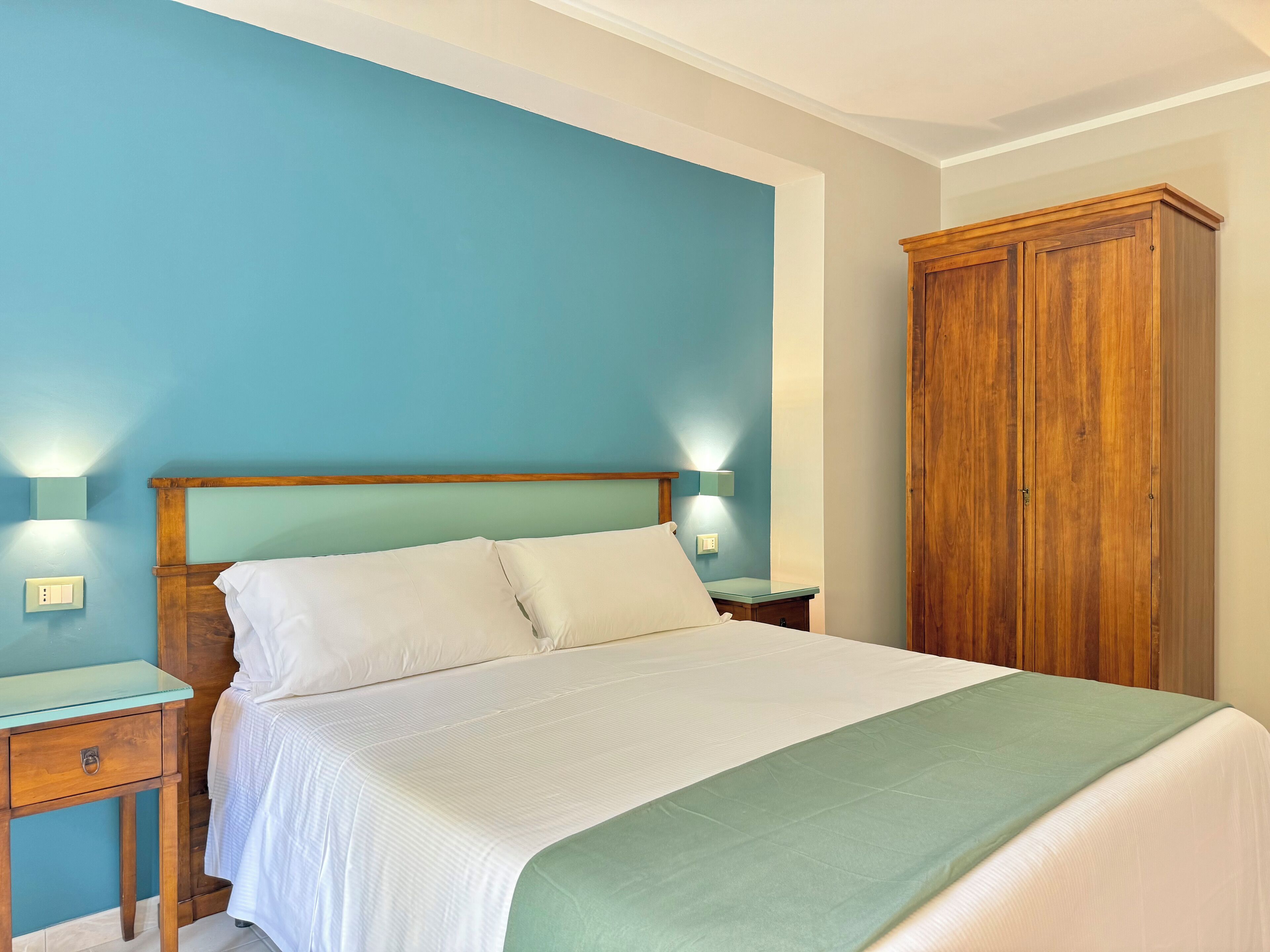 Un dormitorio con una cama, dos mesitas de noche, un armario junto a la cama y un guardarropa de madera.