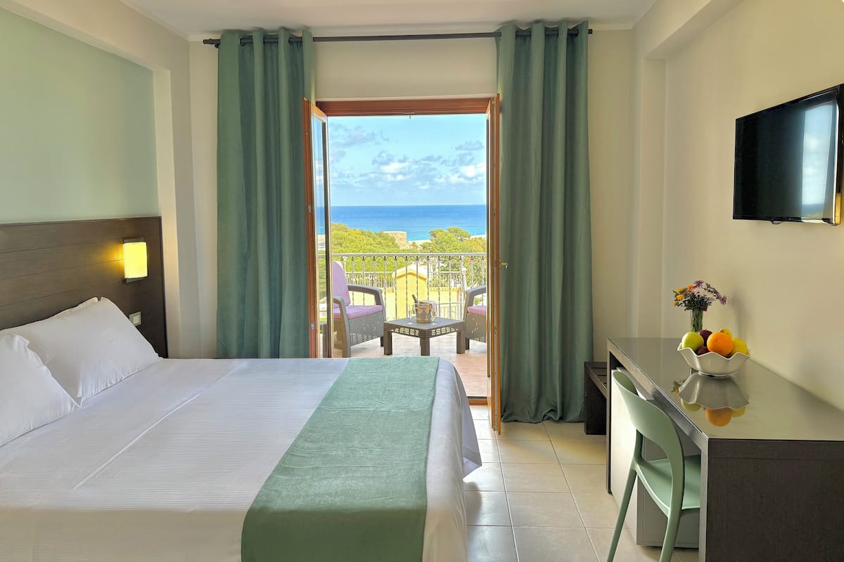 Ein Hotelzimmer mit Bett, Schreibtisch, Fernseher und einem Balkon mit Meerblick.