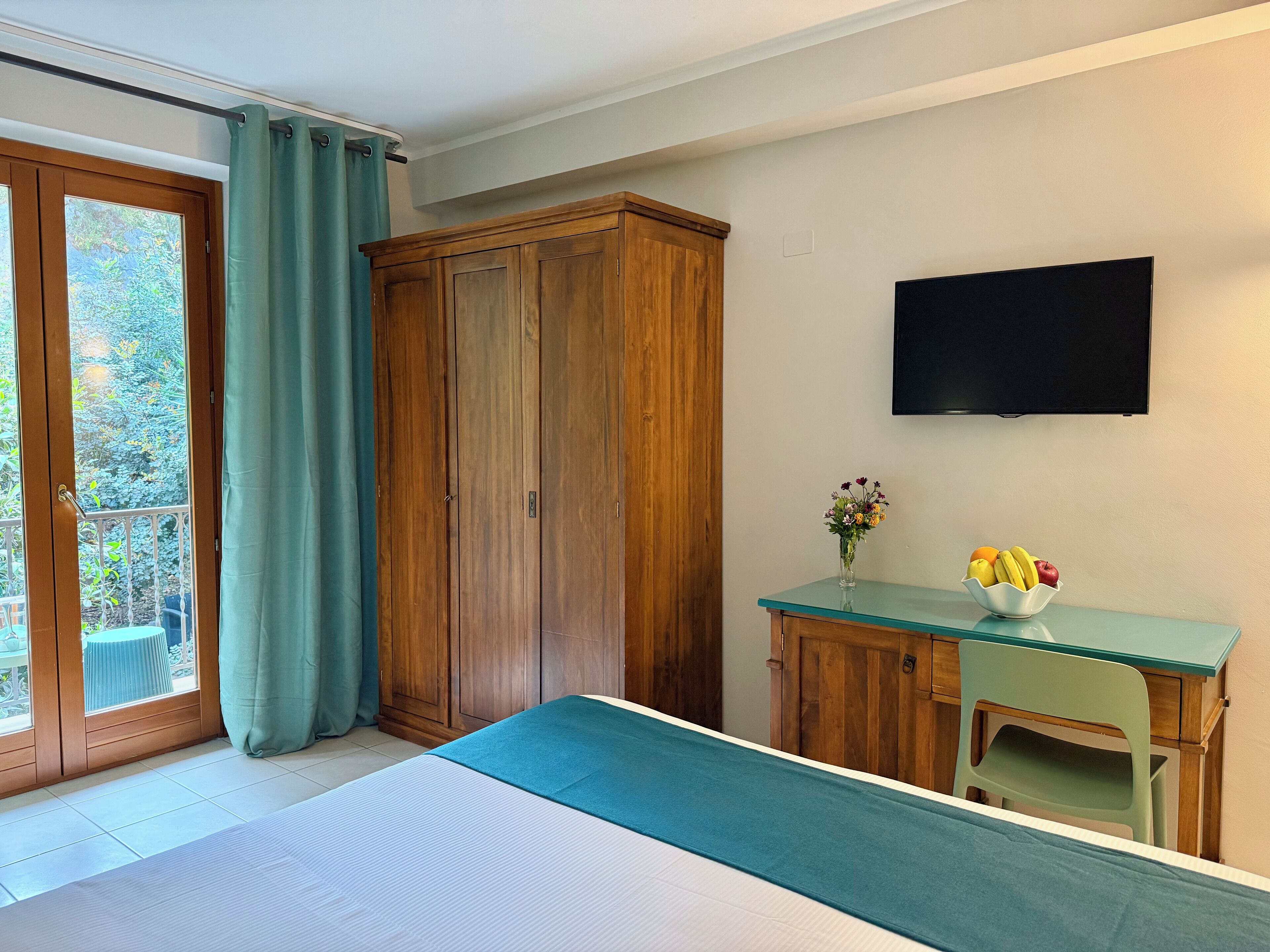 Una camera da letto con un letto, un armadio in legno, una scrivania con un vaso di fiori e una televisione a parete.