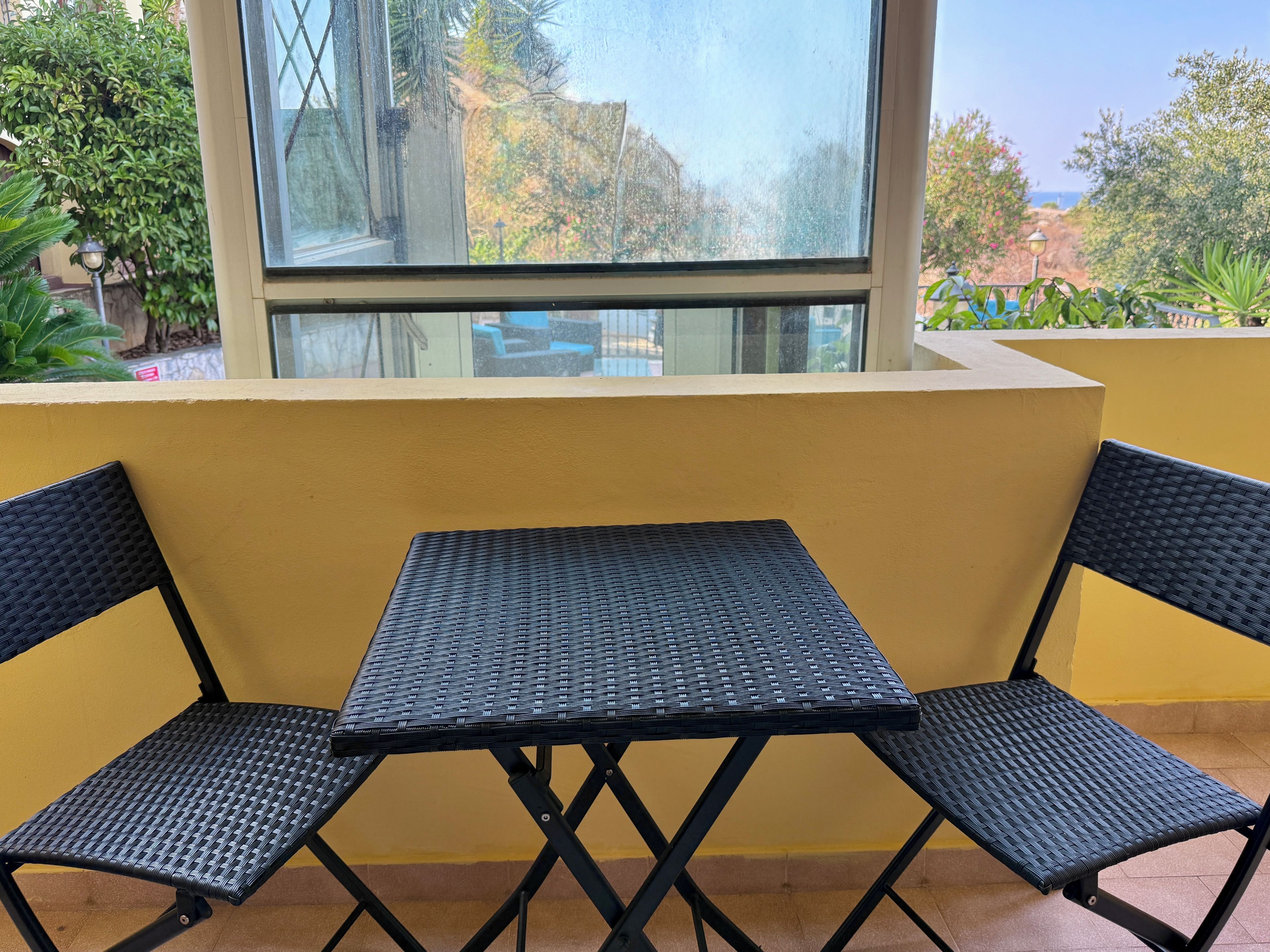 A small outdoor seating area with a table and two chairs, a yellow wall, and a window overlooking a garden.