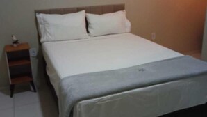 Ein ordentlich bezogenes Bett mit weißen Leinen und einer grauen Decke, ein Nachttisch aus Holz mit einer Lampe und ein an der Wand befestigtes Regal.
