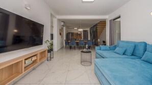 Ein modernes Wohnzimmer mit großem Flachbildfernseher, einem blauen Sofa, einer Holz-Unterhaltungsecke und einem Essbereich mit Tisch und Stühlen.