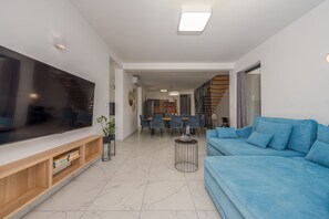 Ein modernes Wohnzimmer mit großem Flachbildfernseher, einem blauen Sofa, einer Holz-Unterhaltungsecke und einem Essbereich mit Tisch und Stühlen.