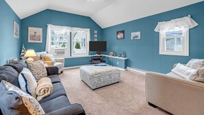A living room with a blue wall, a ceiling fan, a TV, and a window with a view of a house. - Coffee | BBQ Grill | Outdoor Dining | Wifi (Belmar)