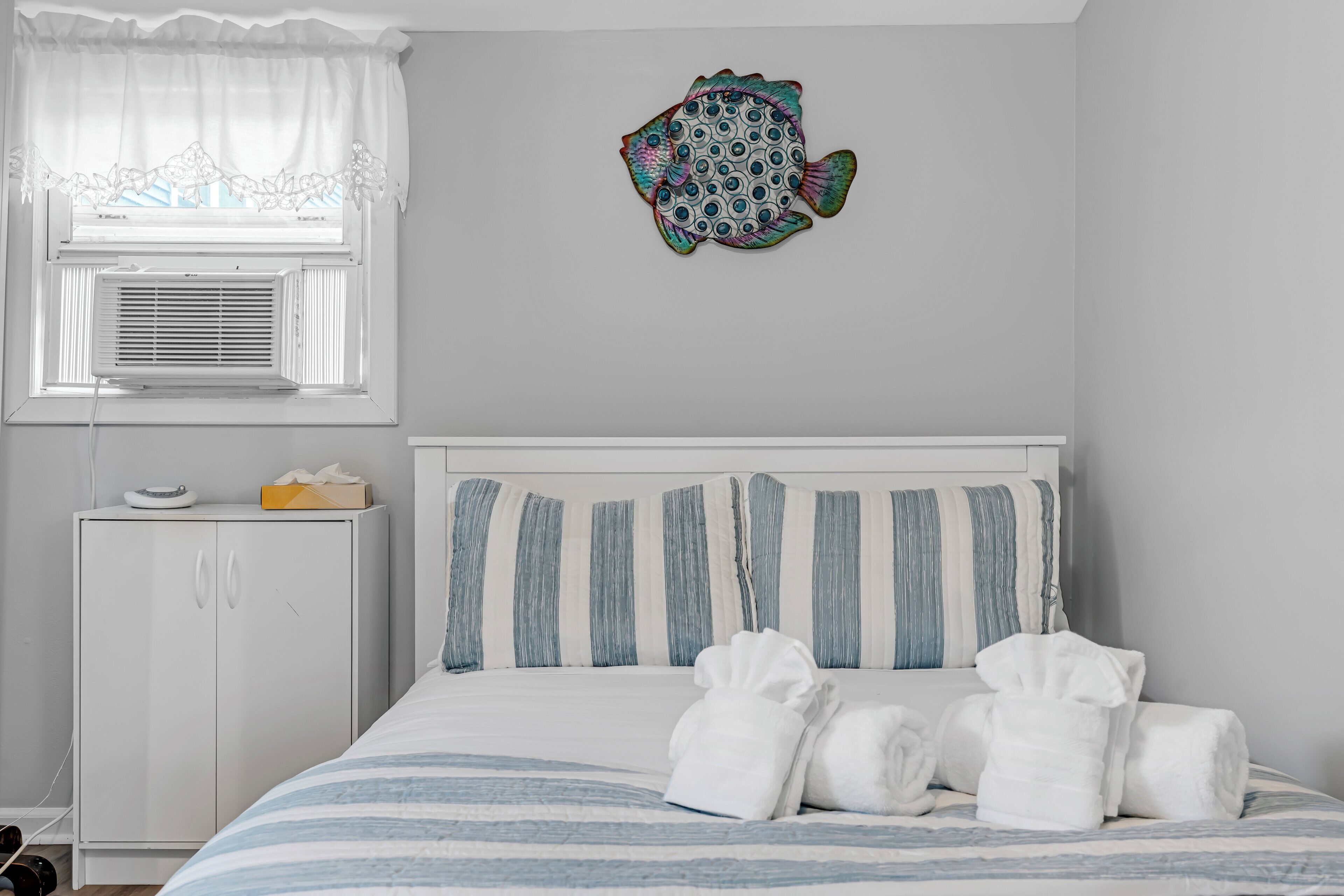 A bedroom with a bed, striped pillows, a fish decoration, a window with curtains, and a white wardrobe.