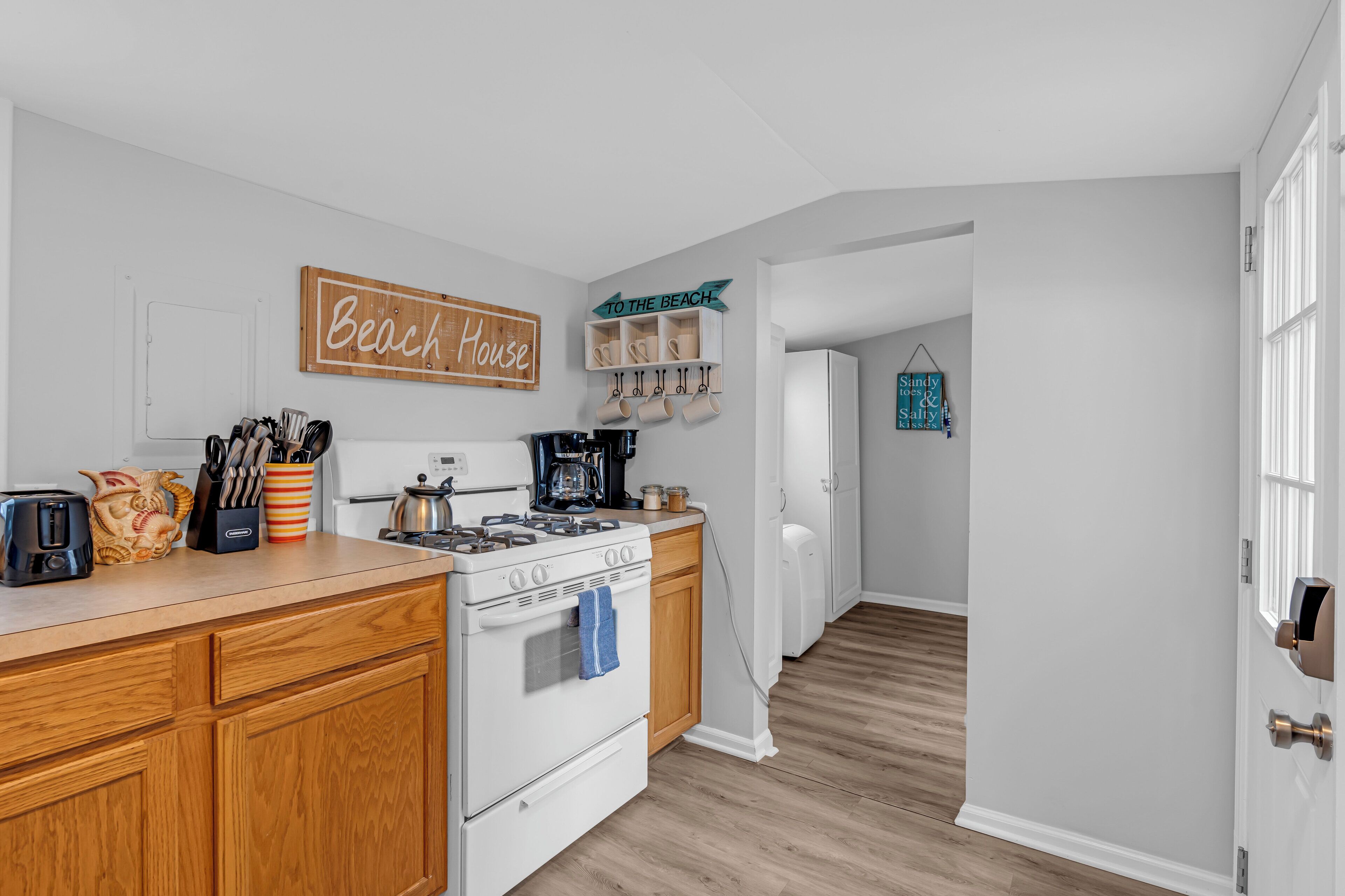 A kitchen with wooden cabinets, a stove, and a coffee maker. There is a sign that reads "Beach House" and another that says "To the Beach."