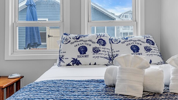 A bedroom with two windows, a bed with a blue and white patterned bedspread, two pillows with 'Ocean View Resort' text, and a bedside table with a phone. - Coffee | BBQ Grill | Outdoor Dining | Wifi (Belmar)