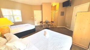 A hotel room with two beds, a small table with chairs, a TV mounted on the wall, and a door leading to another room.