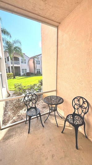 A balcony with two metal chairs and a small table, overlooking a building with a green lawn and palm trees.