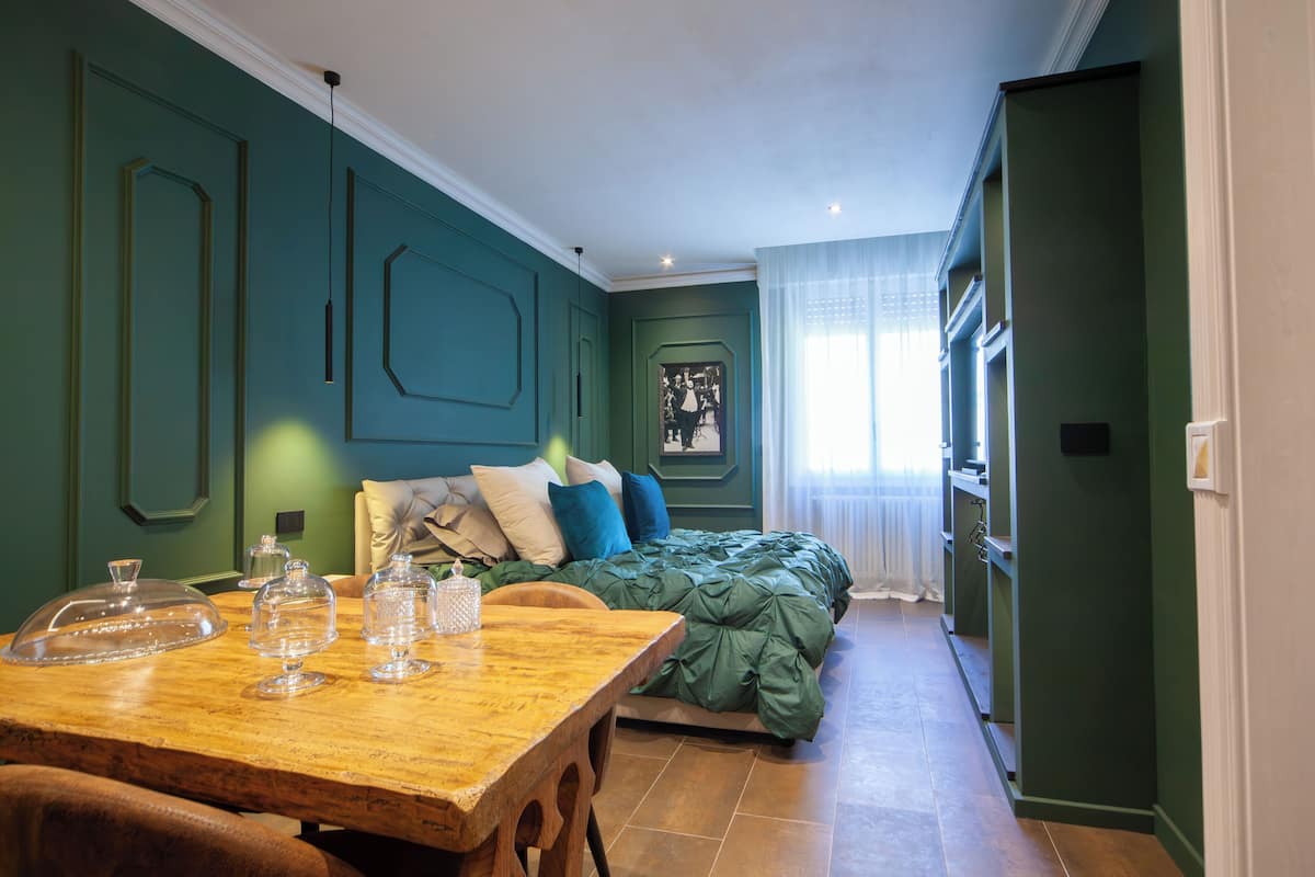 A room with a bed, a wooden table with glassware, and a green wall with a geometric design.